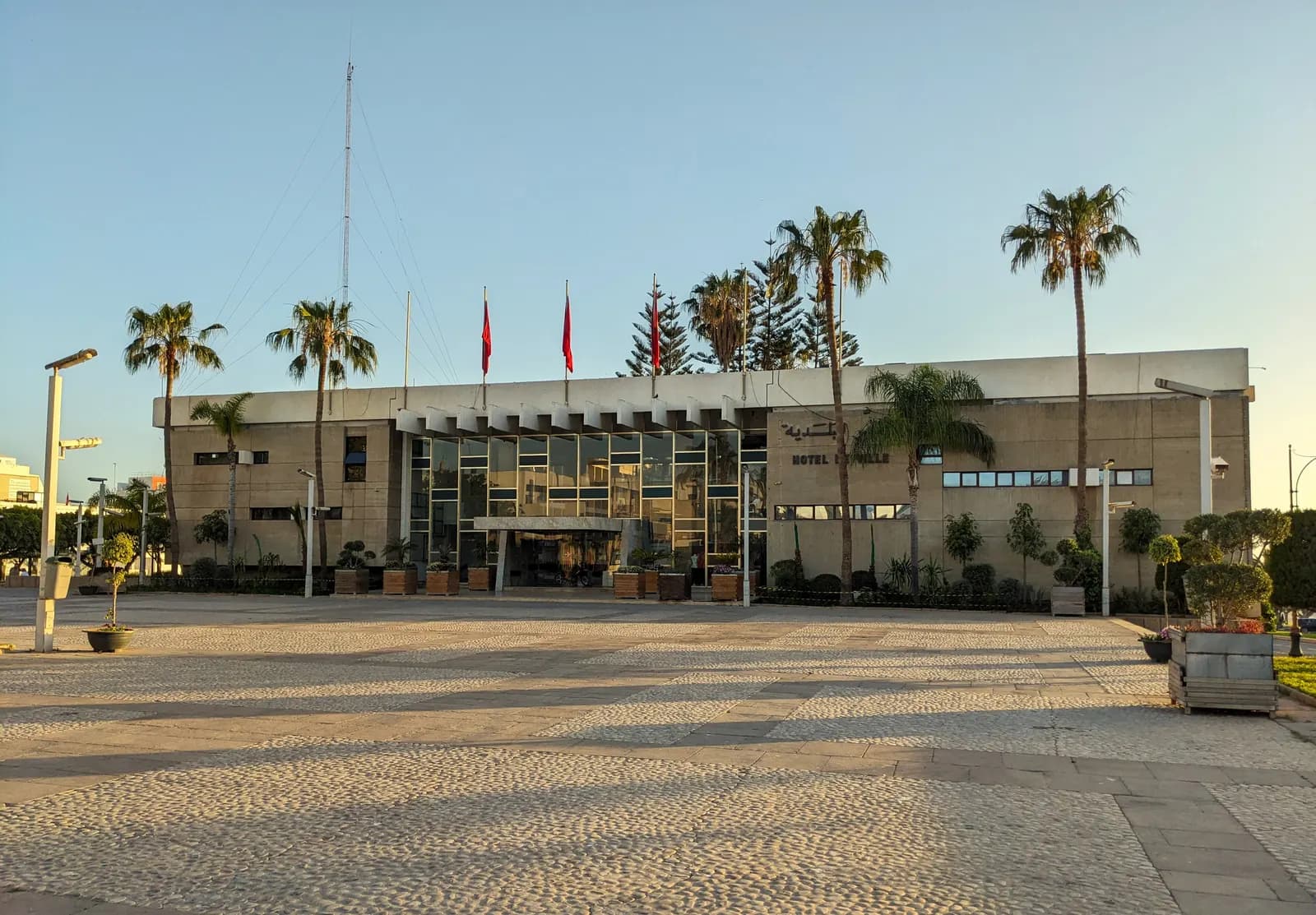 The concrete and glass city hall of Agadir behind a wide paved square, with palm trees and flags in late afternoon light