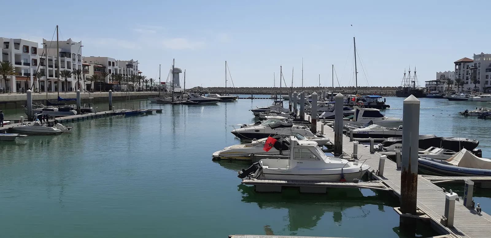 Motor boats and yachts moored on the pontoons of Agadir marina, with white apartment buildings and palm trees along the quay