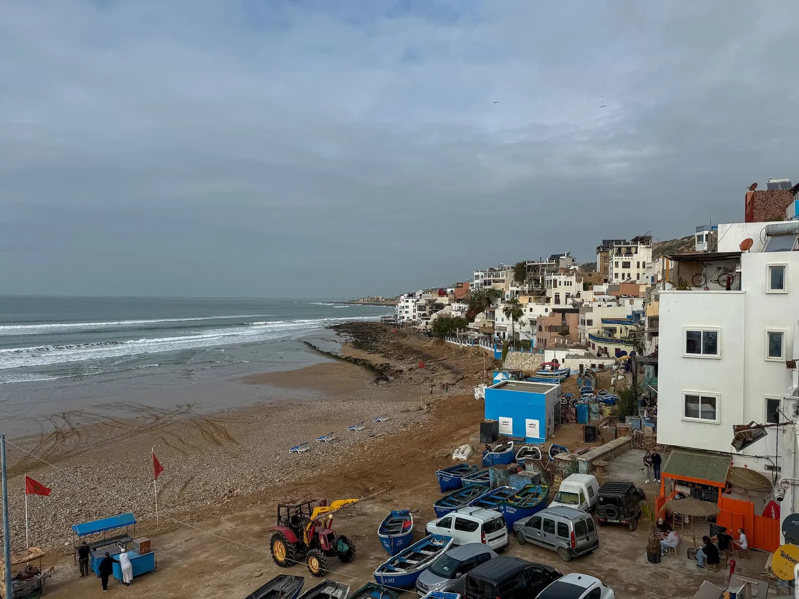 Taghazout village above its beach under a grey sky, with blue fishing boats, a tractor and parked cars on the sand by the houses