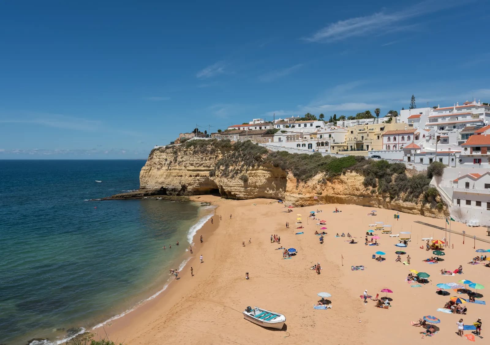 Carvoeiro beach in a small cove below white houses and golden cliffs, with sunshades, a fishing boat on the sand and blue sea