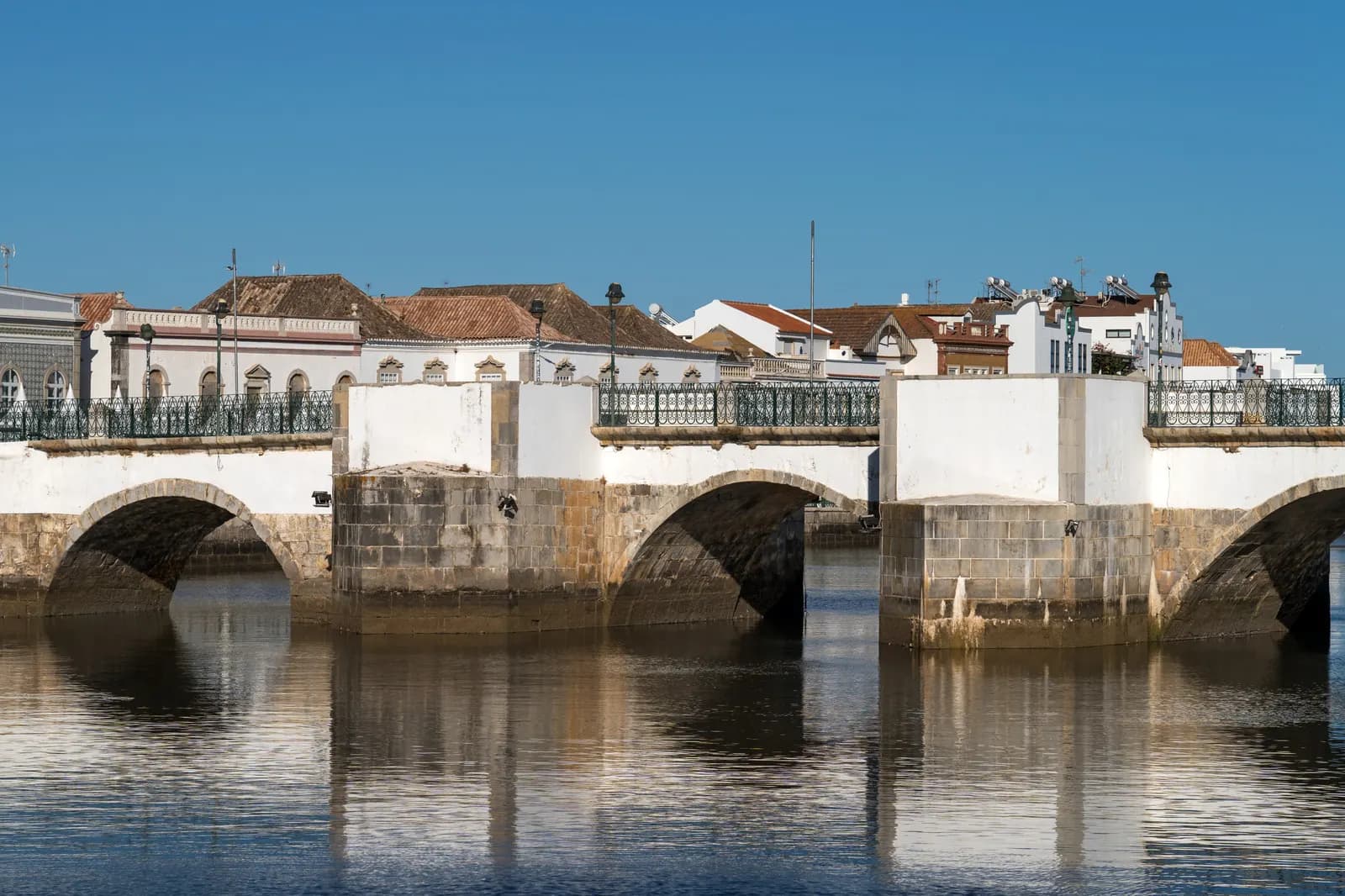 The arches of the old bridge over the Gilão river in Tavira reflected in the water, with white houses behind