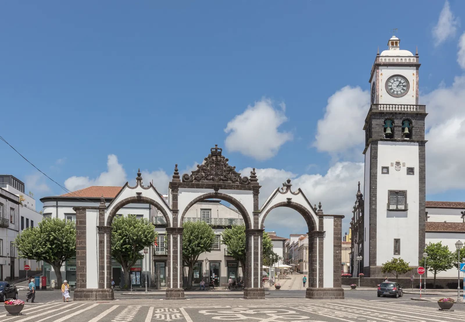 The three stone arches of the Portas da Cidade in Ponta Delgada with the white clock tower beside them, on a square paved in black and white