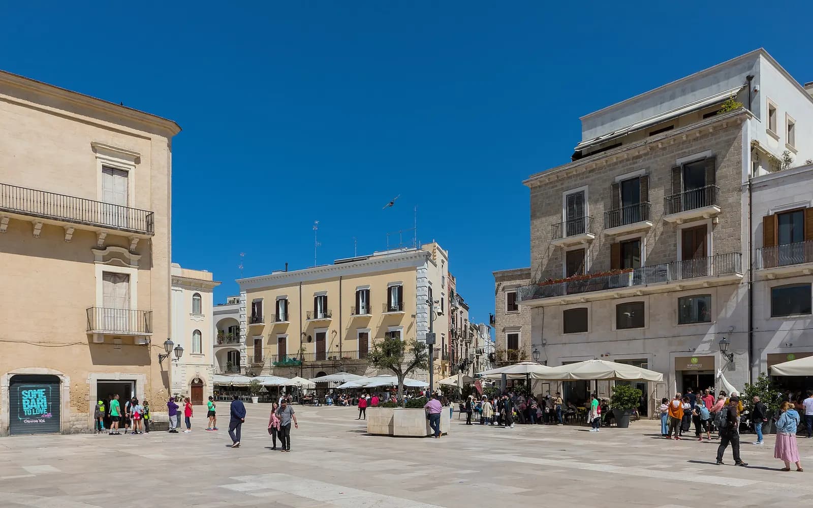 Piazza Mercantile in Bari Vecchia, a paved square lined with pale stone buildings and cafe umbrellas, with people walking