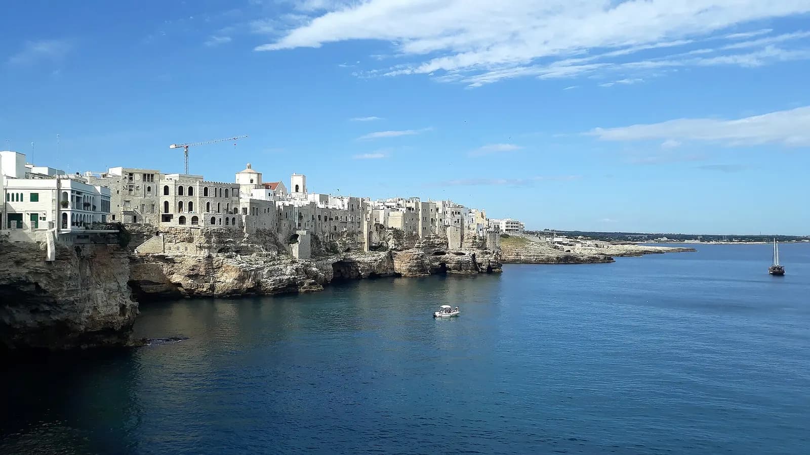 The white houses of Polignano a Mare along the top of limestone cliffs above a calm blue sea, with a small boat in the bay