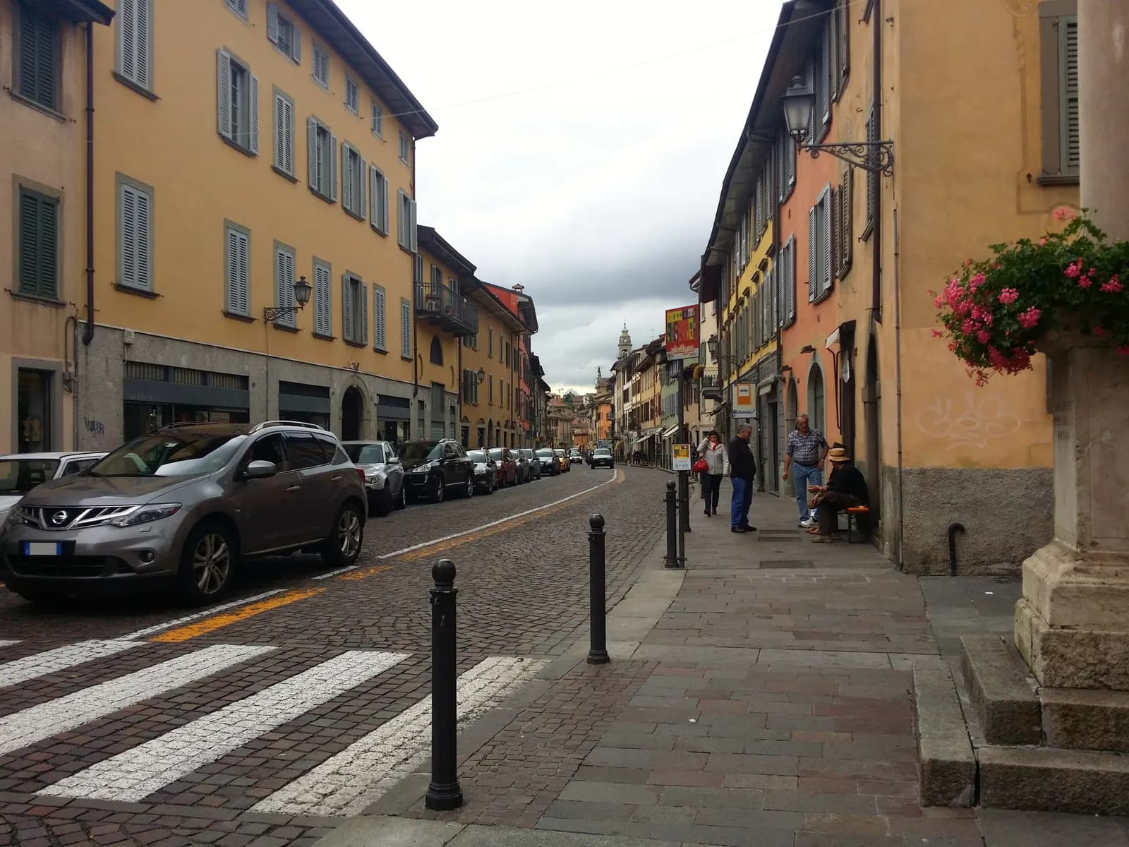 Via Santa Caterina, the cobbled main street of Borgo Santa Caterina, lined with ochre and pink houses, small shops and parked cars under a cloudy sky.