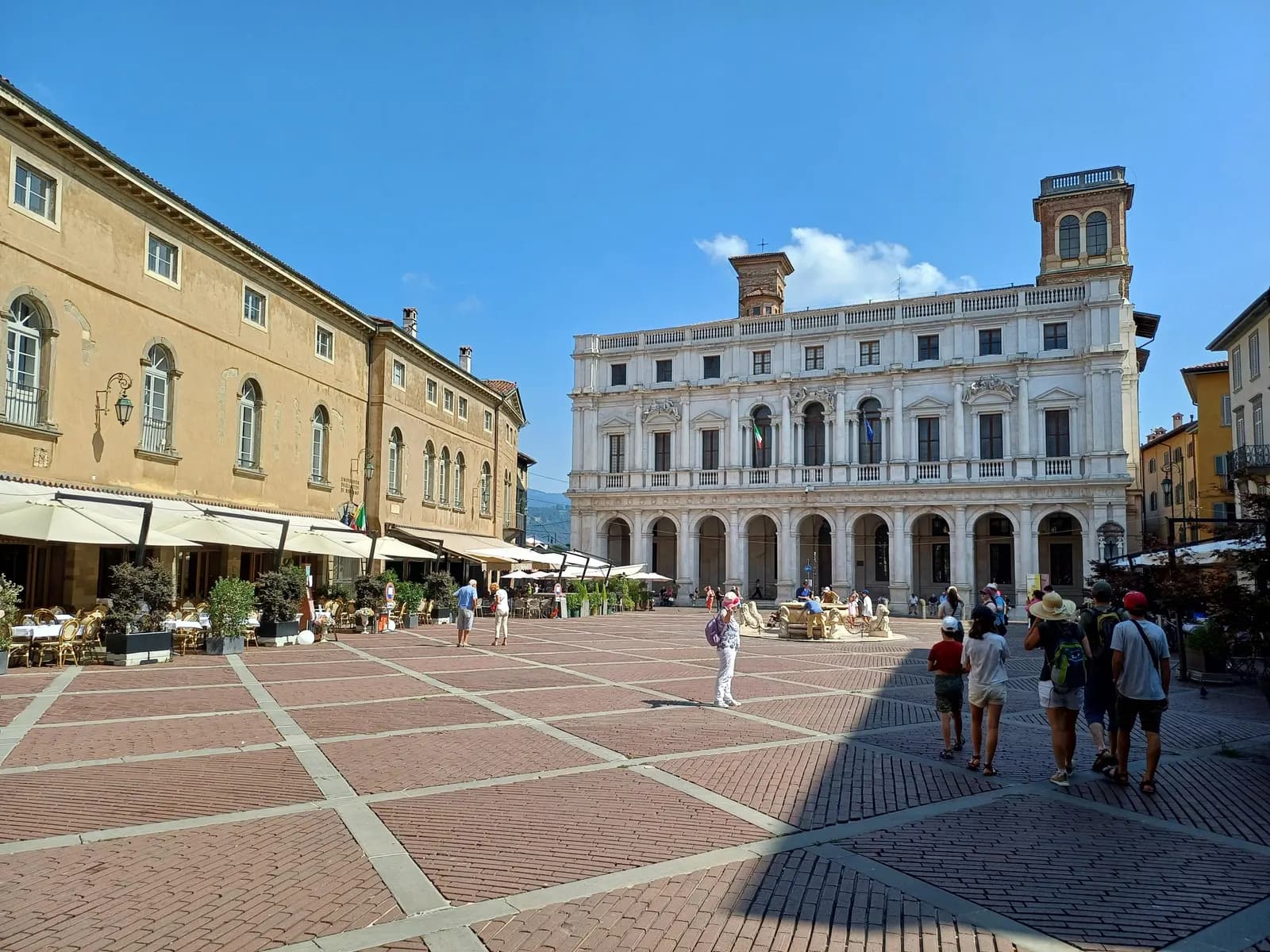 Piazza Vecchia in Città Alta on a sunny day, with café terraces on the left and the white arcaded Palazzo Nuovo and the Contarini fountain ahead.