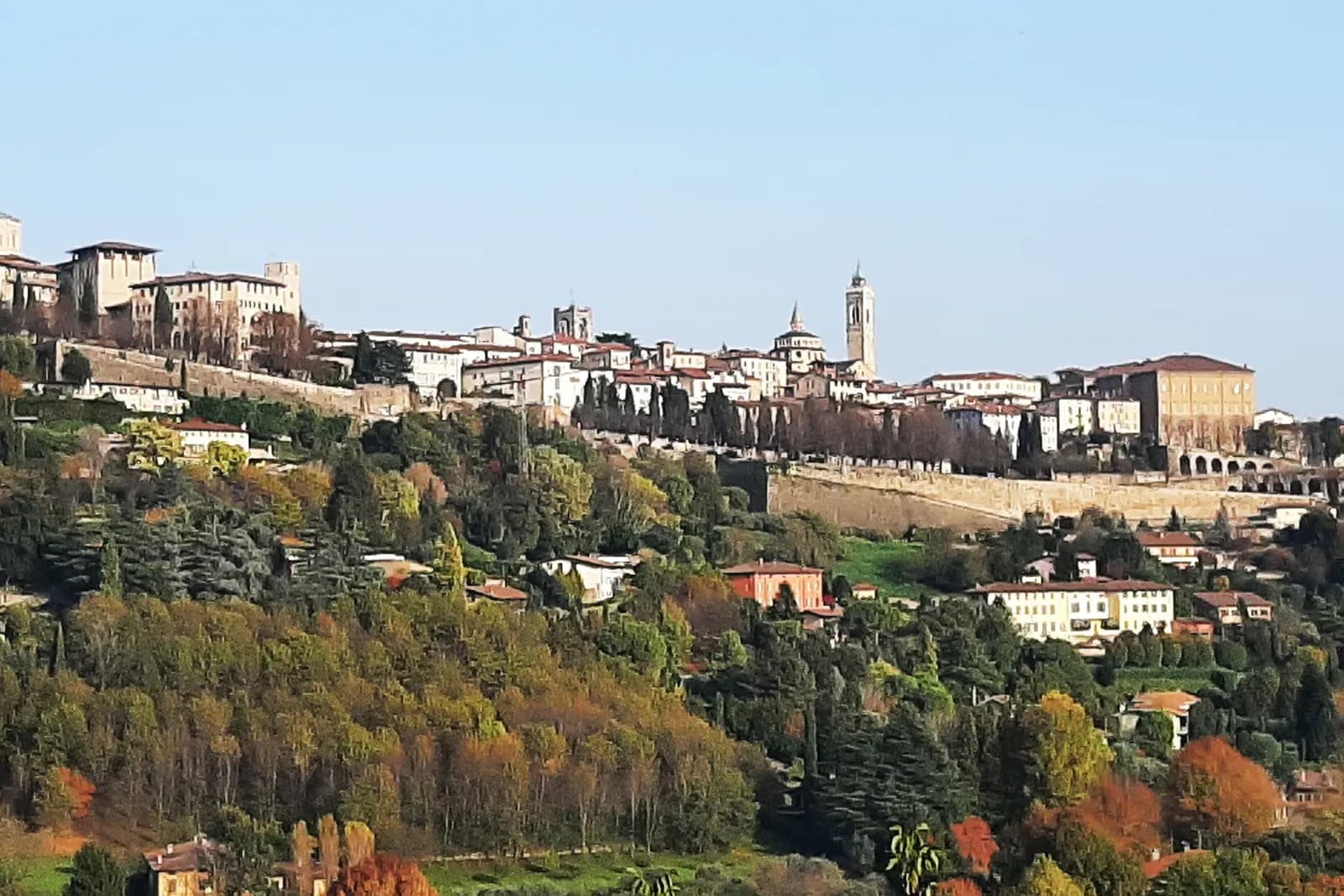 Città Alta on its hilltop in autumn, a line of pale houses, domes and bell towers above the Venetian walls and wooded slopes.