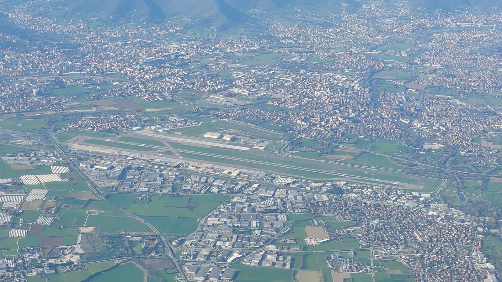 Orio al Serio airport seen from a plane, the runway and terminal between warehouses, fields and houses, with hills in the distance.
