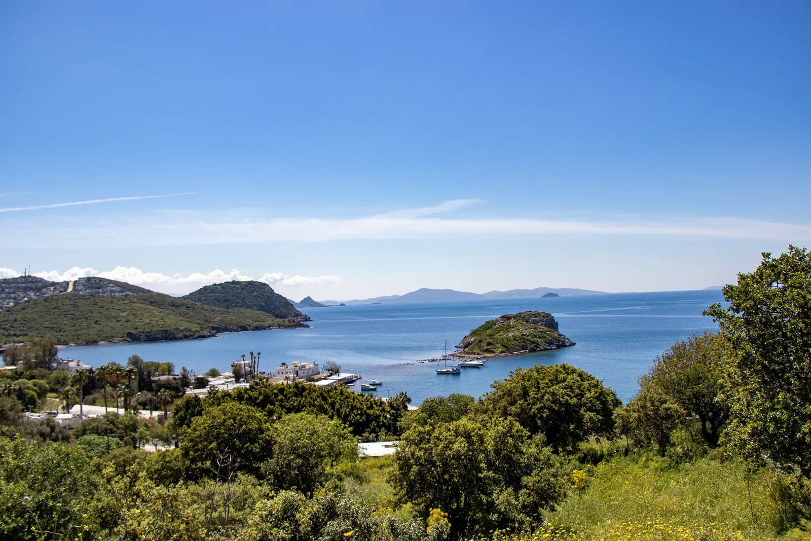 Gümüşlük bay seen from a green hillside, with Rabbit Island near the shore, boats at anchor and islands on the horizon