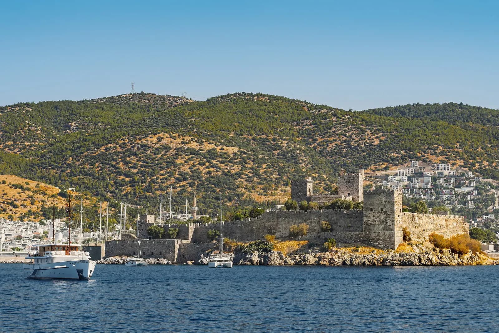 The walls and towers of Bodrum Castle on its rocky point, with a motor yacht in front and white houses on the pine-covered hills behind