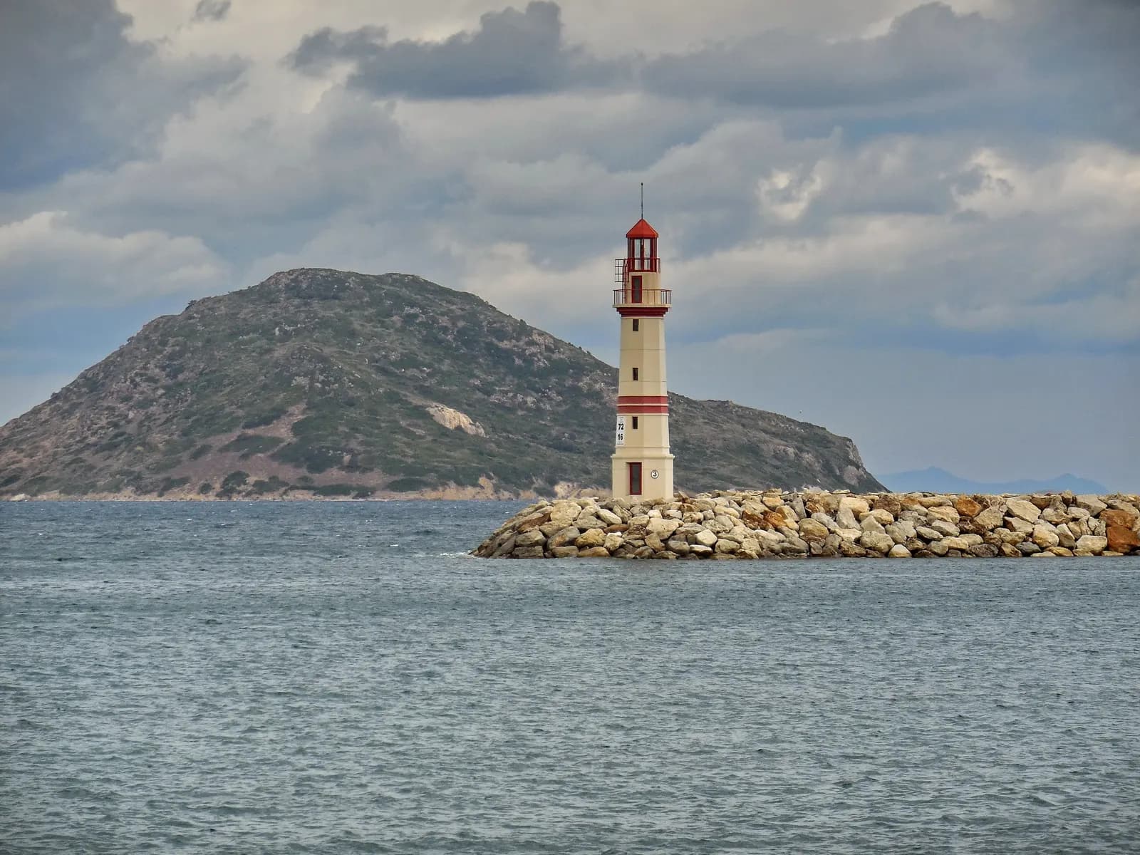 A cream and red lighthouse on the rock breakwater at Turgutreis, with a hilly island behind under a cloudy sky