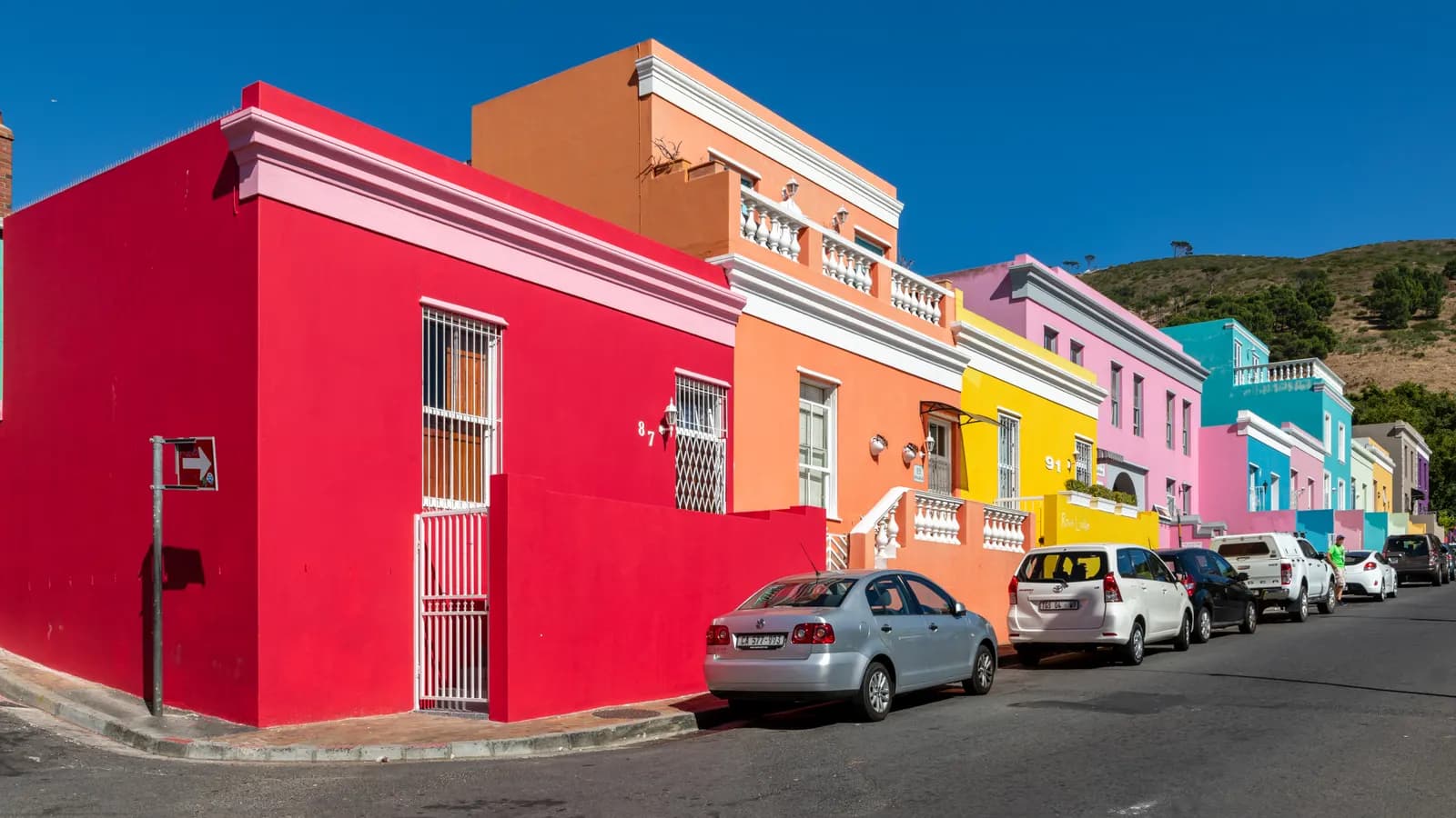 Houses painted red, orange, yellow, pink and blue along Wale Street in Bo-Kaap, with cars parked at the kerb and a hill behind