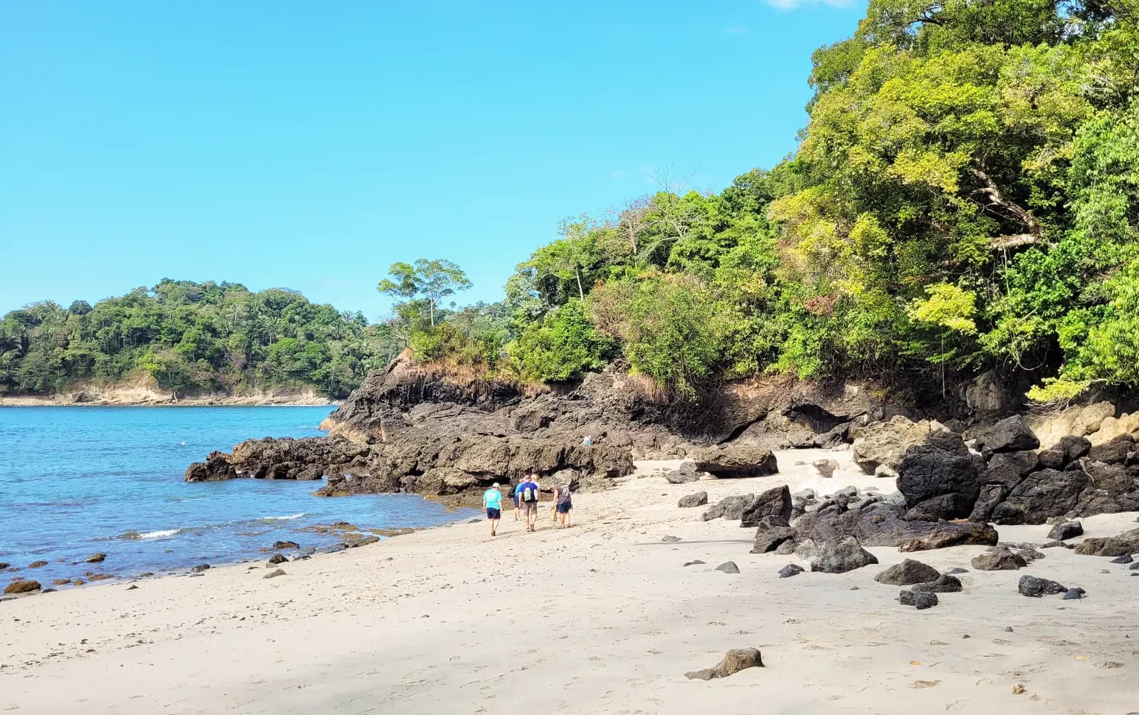 Walkers on a pale sand beach in Manuel Antonio National Park, with dark rocks, forest-covered headlands and blue sea