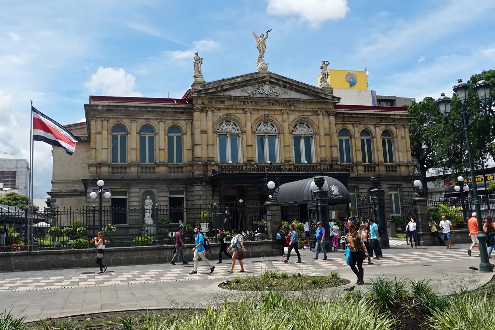 The stone facade of the National Theatre in San José, with statues on the roof, a Costa Rican flag and people walking across the square in front