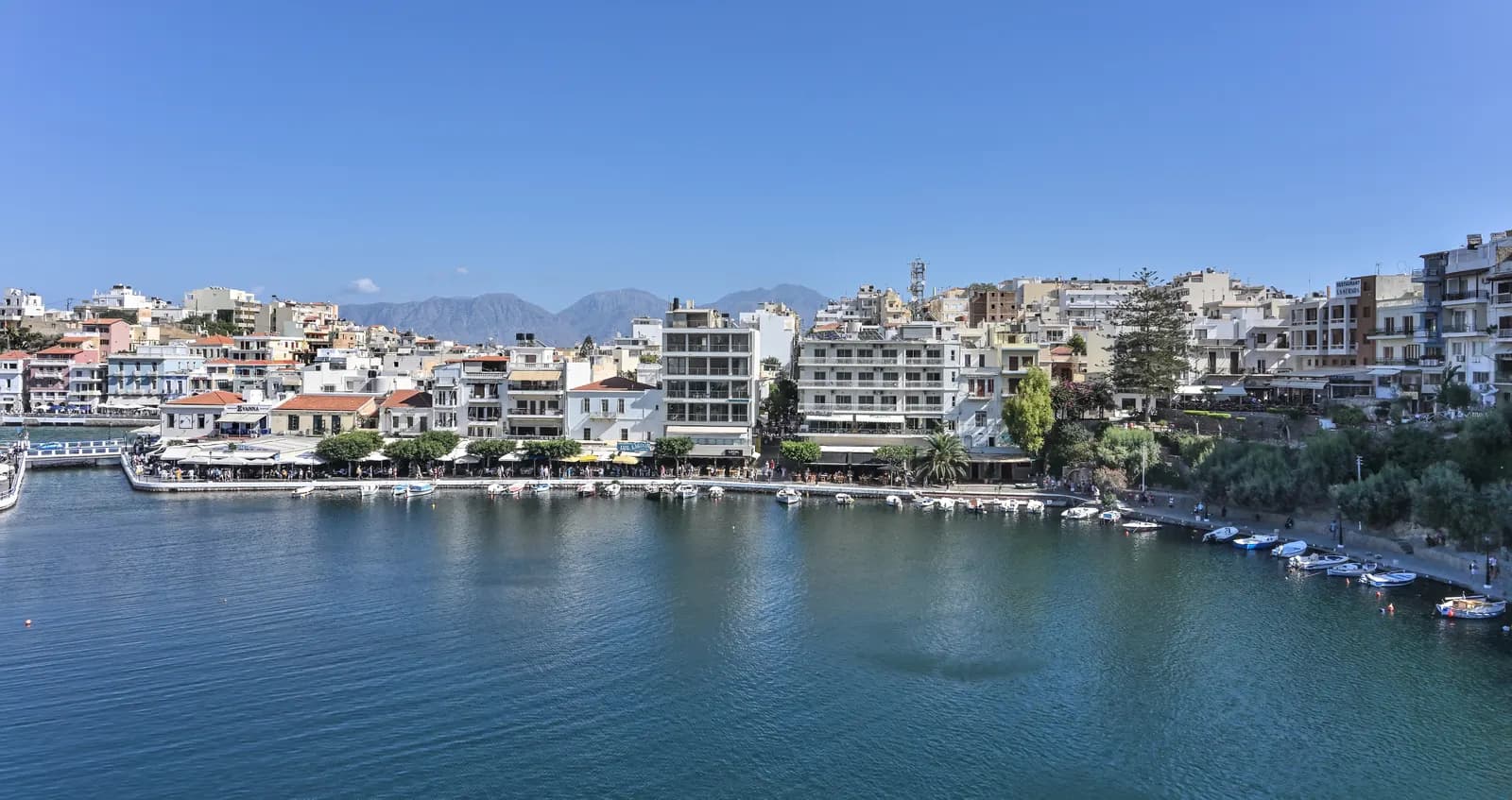 Lake Voulismeni in Agios Nikolaos ringed by cafes and apartment buildings, with small boats along the quay and mountains behind