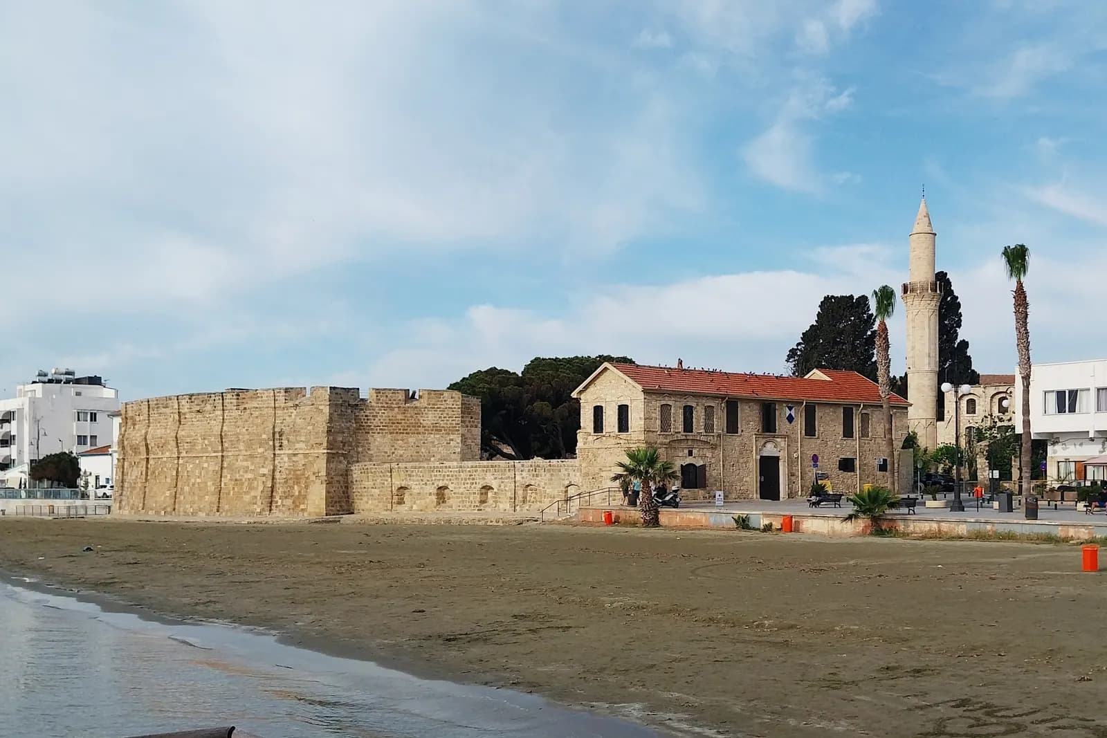 The stone walls of Larnaca's medieval castle and its red-roofed building on the town beach, with palm trees and a minaret behind under a partly cloudy sky