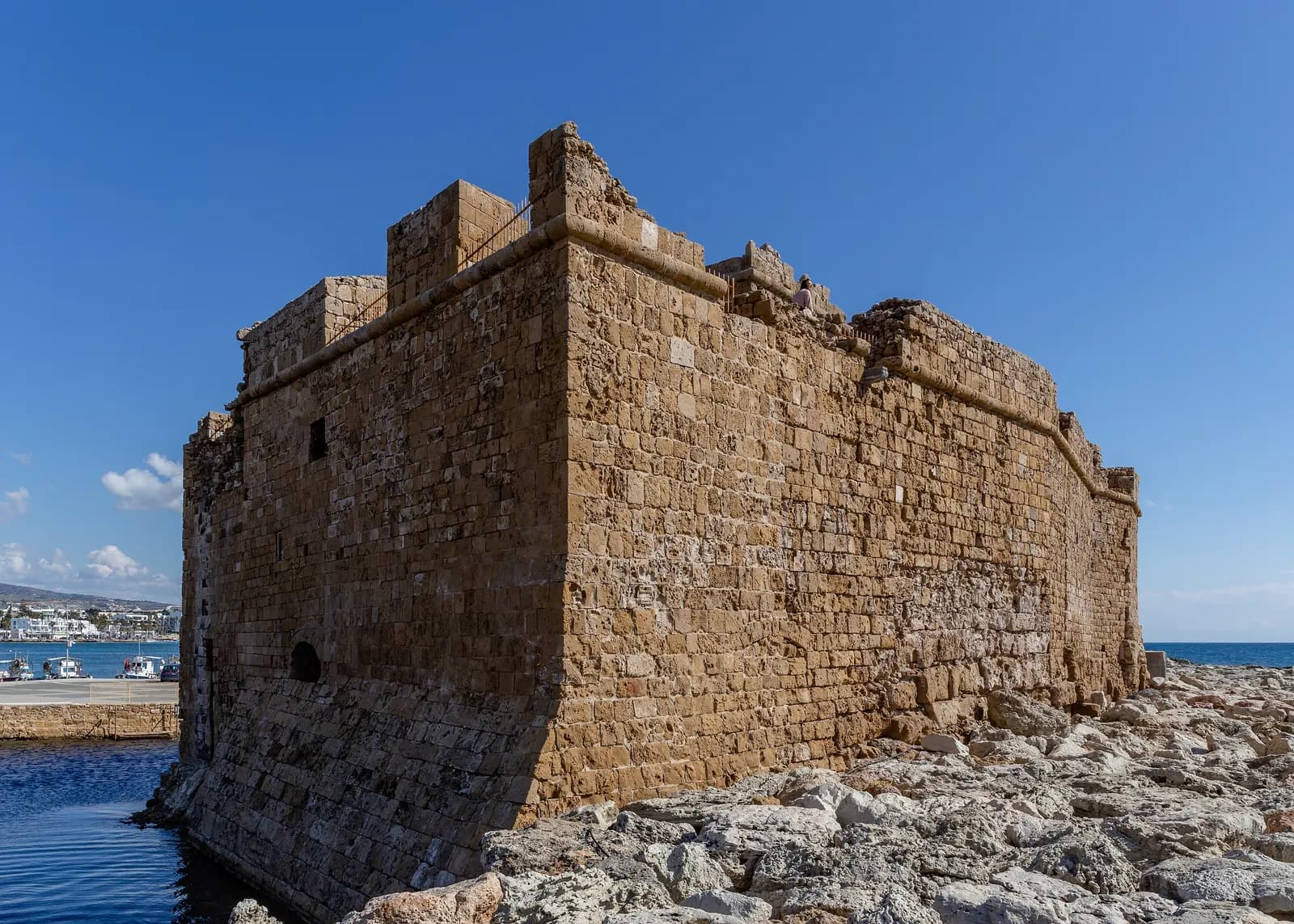 The stone walls of Paphos fort at the end of the harbour under a clear blue sky, with moored boats and the seafront of Kato Paphos behind on the left