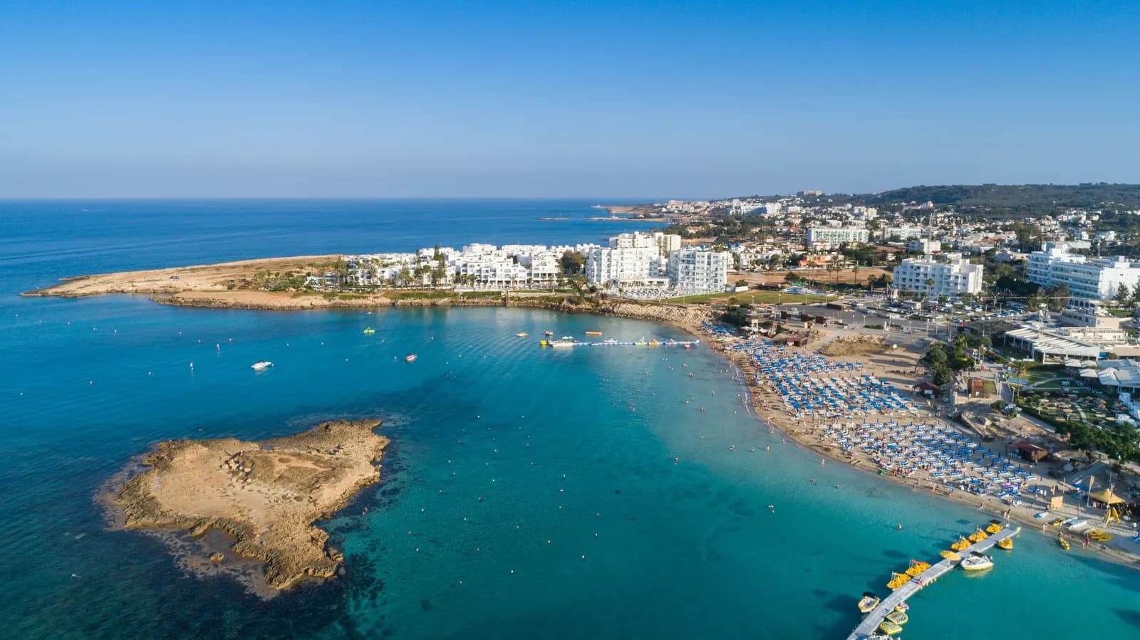Aerial view of Fig Tree Bay in Protaras, with a small rocky islet in turquoise water, rows of beach umbrellas along the sand and hotels behind