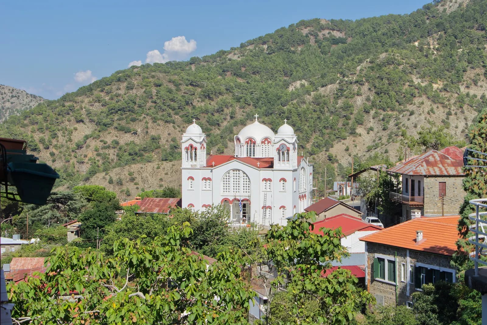 A large white church with domes among the red roofs of Pedoulas, a village in the Troodos mountains, with pine-covered slopes behind under a blue sky