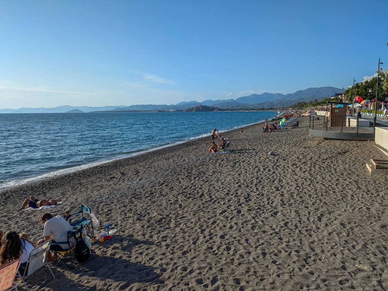Çalış beach with people on the dark sand, the promenade on the right and mountains across Fethiye bay