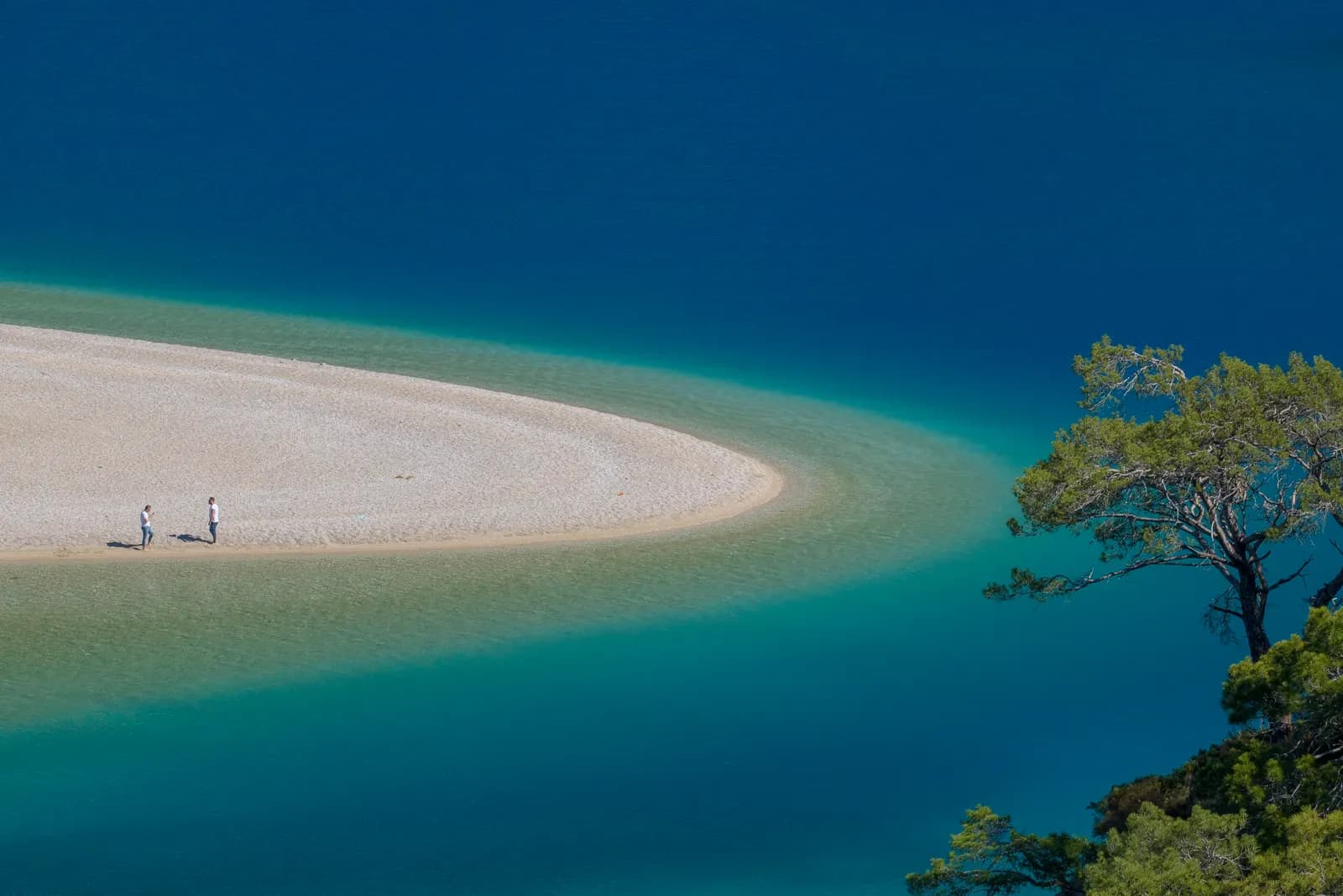 Two people on the white sand spit at the mouth of the Ölüdeniz lagoon, with shallow turquoise water and a pine tree on the shore