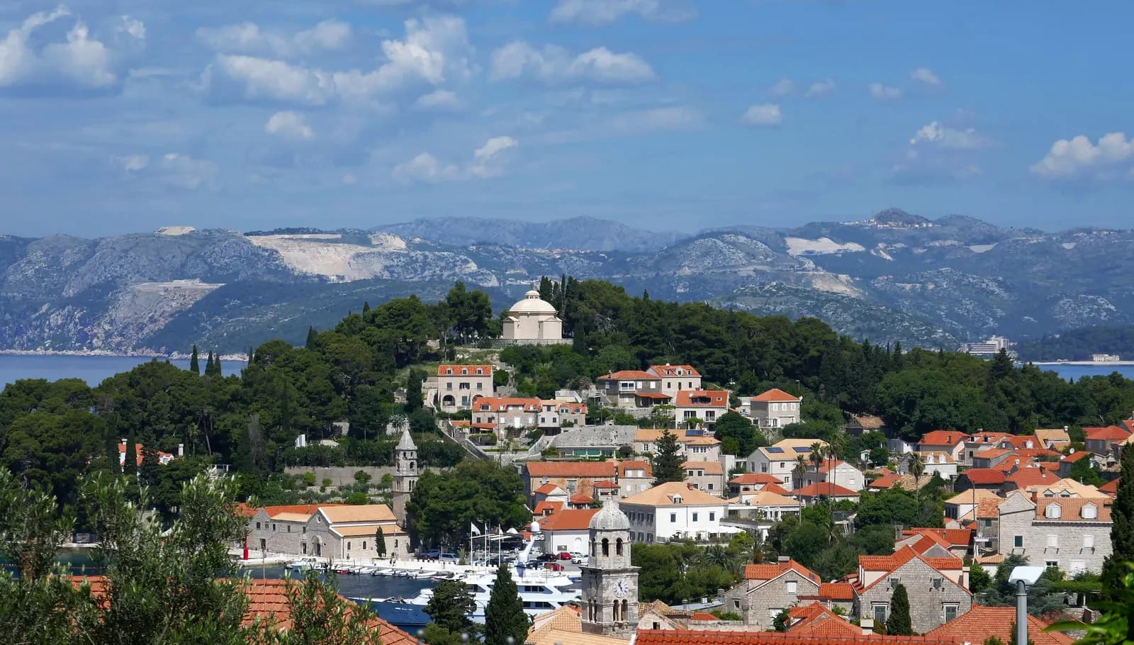 Cavtat's stone houses and harbour below its wooded peninsula, with the white domed mausoleum on the crest and mountains across the bay