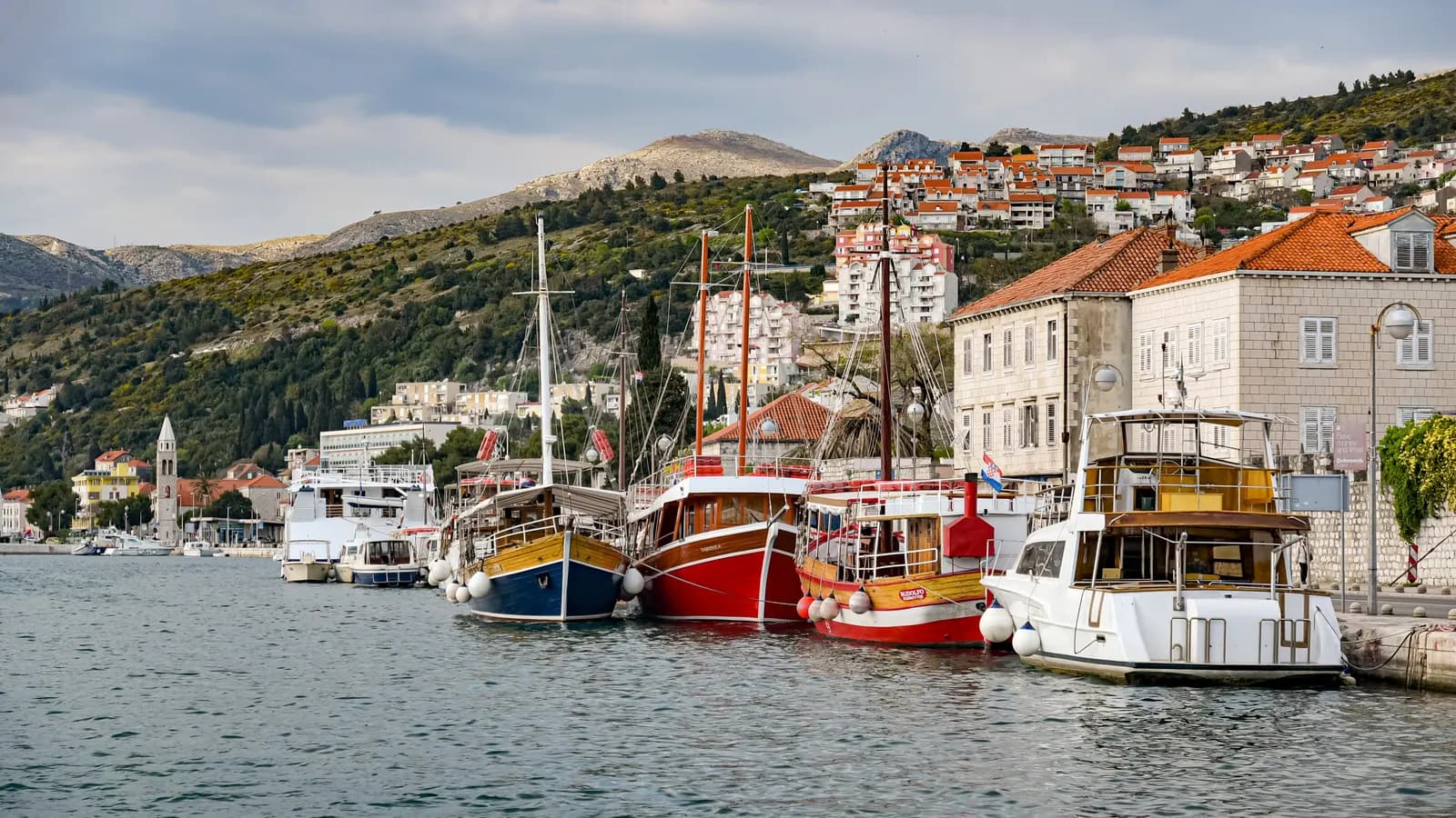 Wooden excursion boats moored along the Gruž waterfront in Dubrovnik, with stone houses and hillside apartment blocks behind