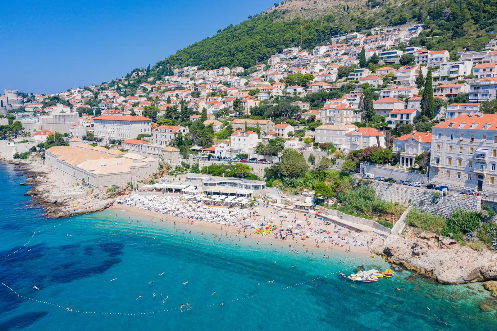 Banje beach below the Ploče district east of Dubrovnik Old Town, with the stone Lazaretto buildings, hillside houses and the city walls in the distance
