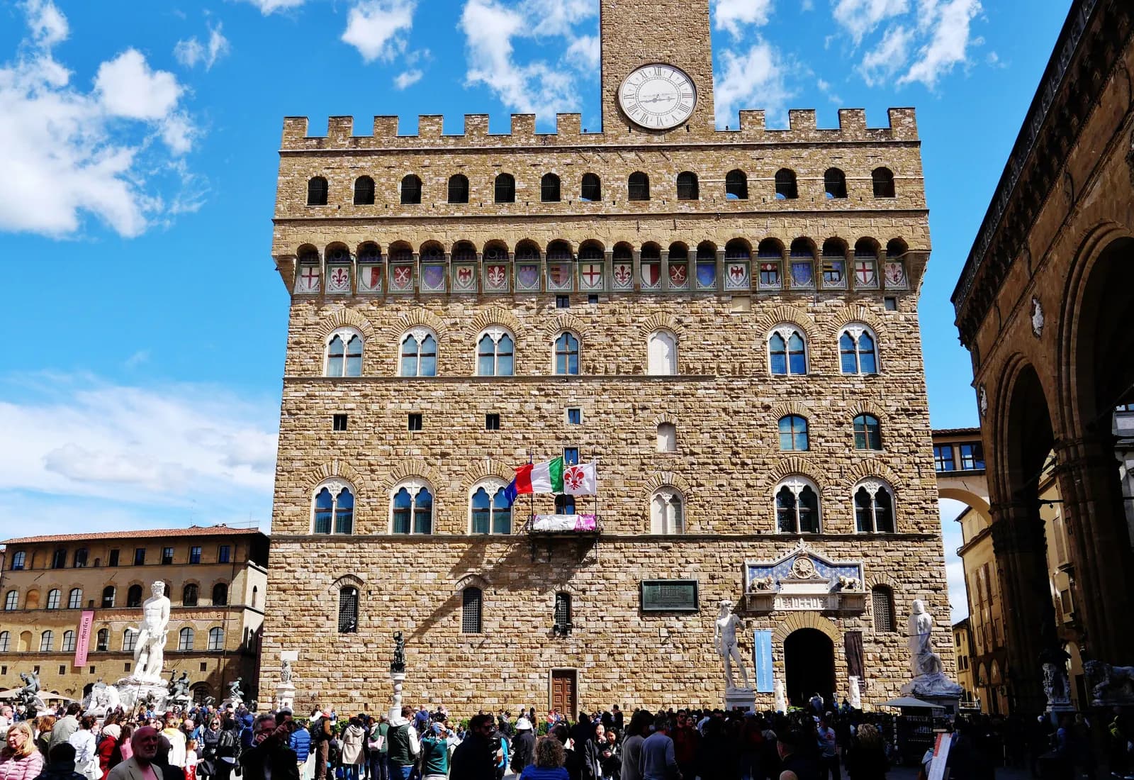 The stone front of the Palazzo Vecchio on Piazza della Signoria, with its clock tower, crowds and the statues by the entrance.