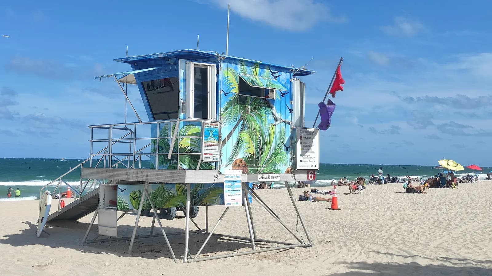 A lifeguard stand painted with palm trees on the sand of Fort Lauderdale beach, with red and purple flags flying, beachgoers under umbrellas and the sea behind