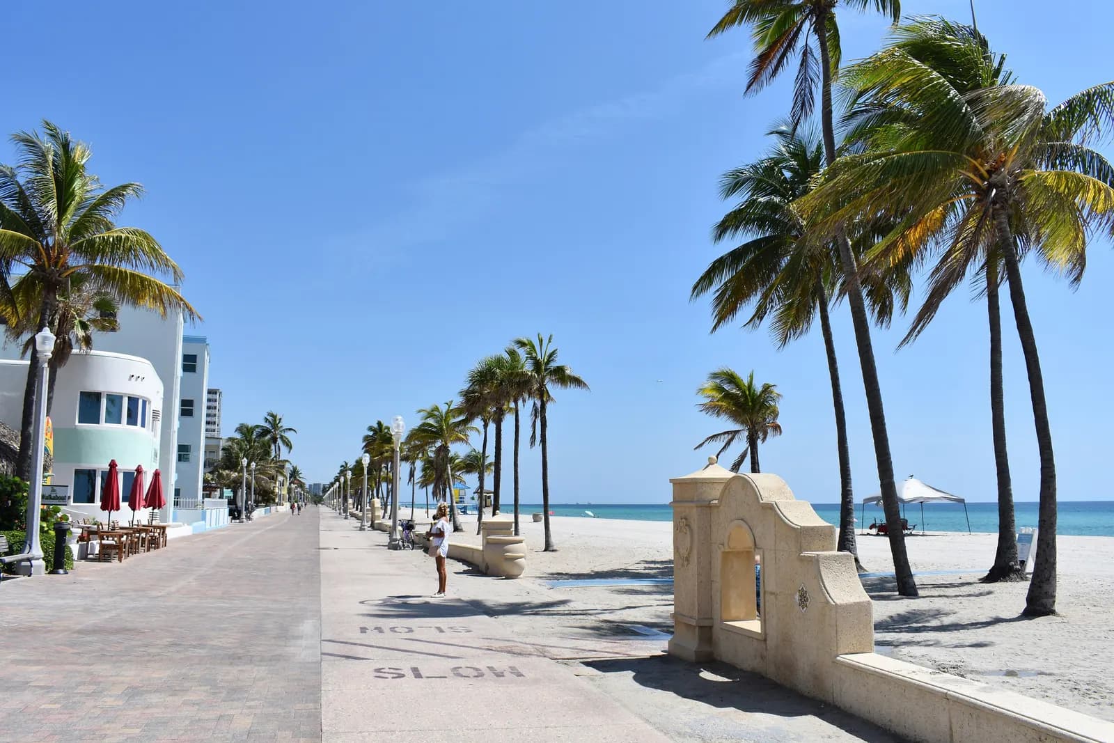 The paved Hollywood Beach Broadwalk lined with palm trees and lamp posts, with a low white building and café tables on the left and the beach and sea on the right