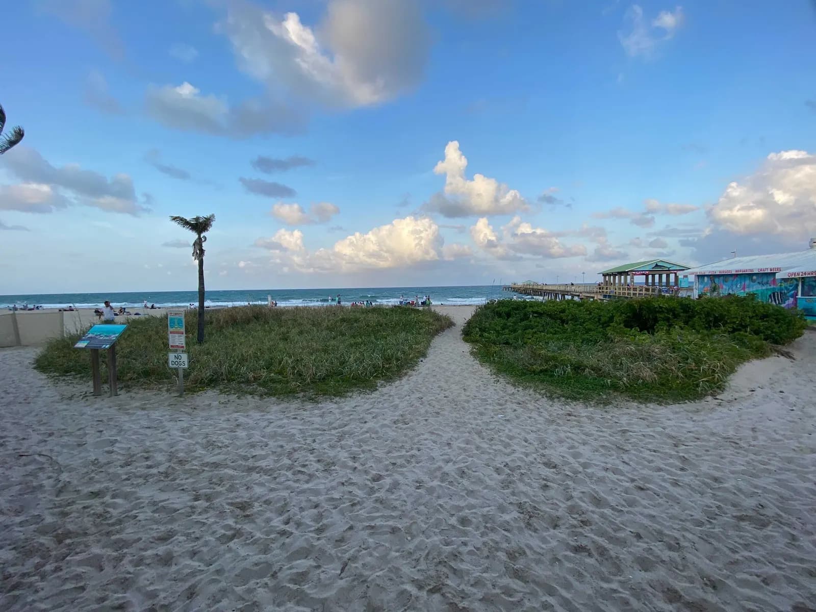 A sandy path between low dune plants leading to the beach at Lauderdale-by-the-Sea, with the fishing pier and a painted beachfront building on the right and the Atlantic under a partly cloudy sky