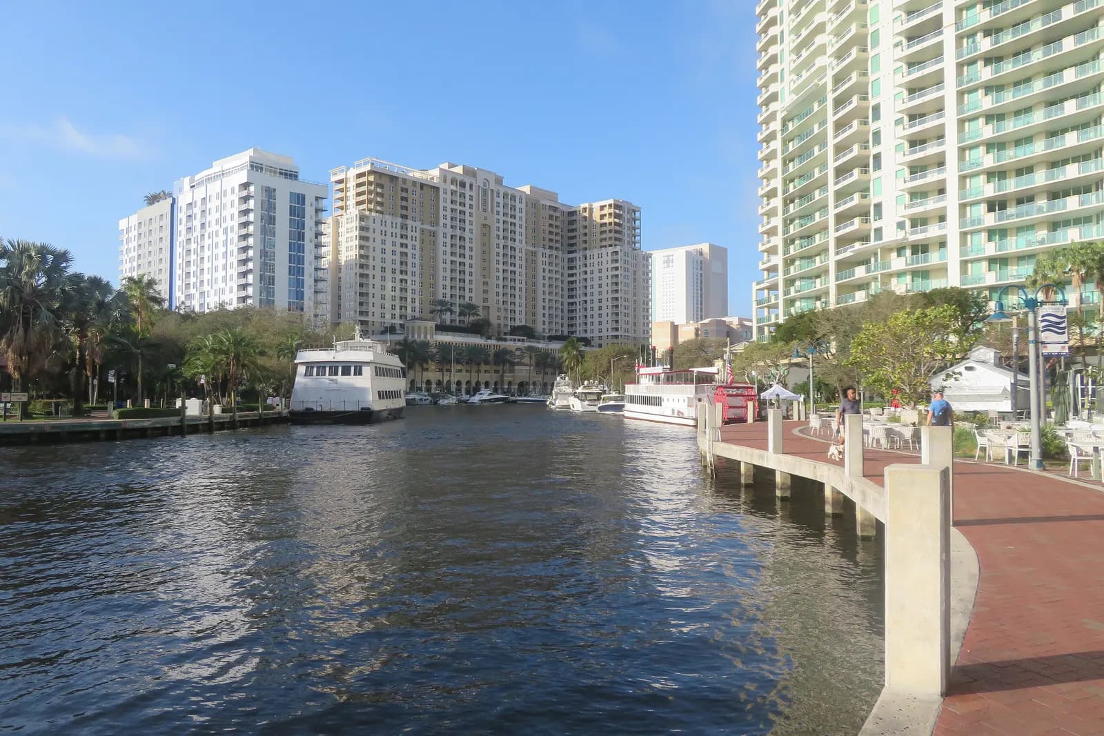 The brick Riverwalk curving along the New River in downtown Fort Lauderdale, with moored boats, palm trees and residential towers under a blue sky
