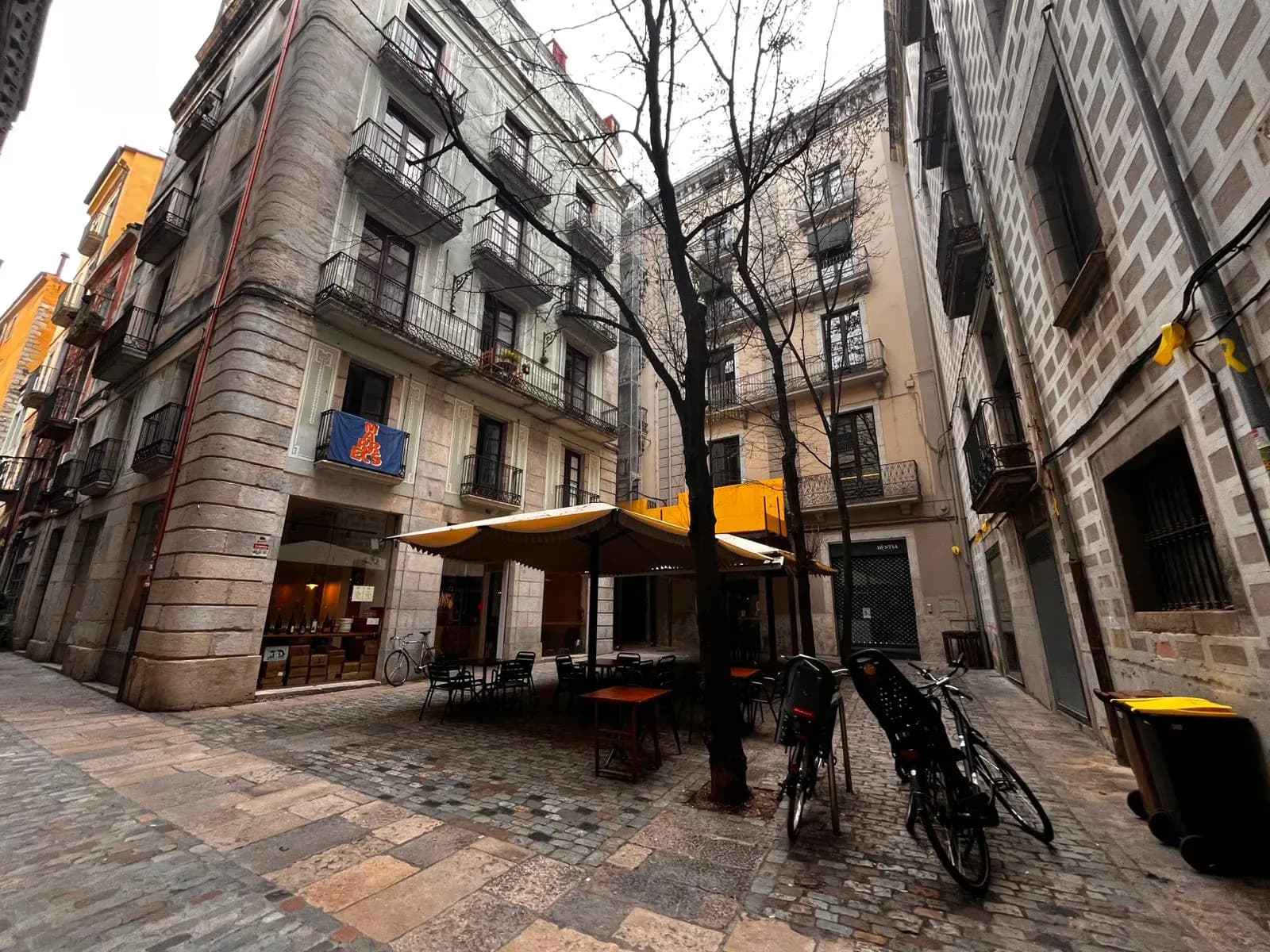 Plaça Bell-Lloc in Girona's Barri Vell, a small stone-paved square with a café terrace, parked bicycles and tall stone houses.
