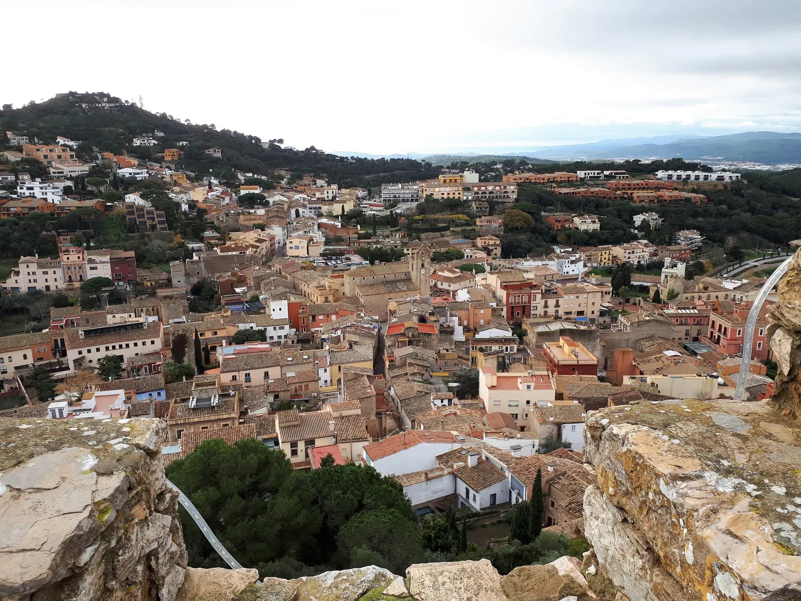 The hill town of Begur seen from its castle, a cluster of tiled roofs around the church, with green hills behind.