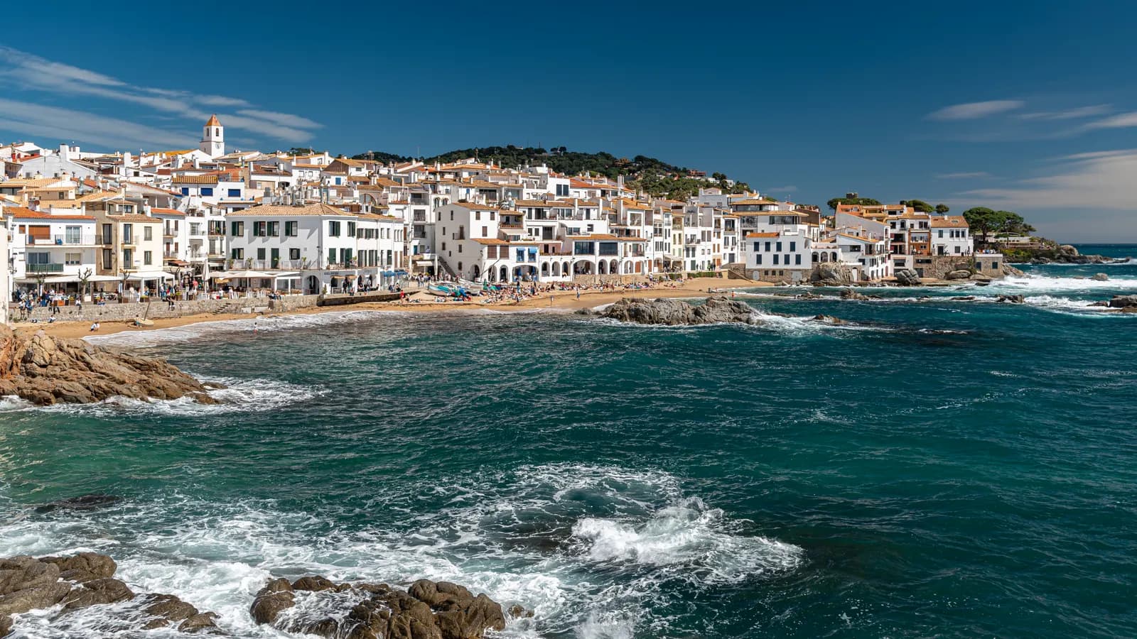 The white seafront houses of Calella de Palafrugell above a small beach, with waves breaking on the rocks in the foreground.
