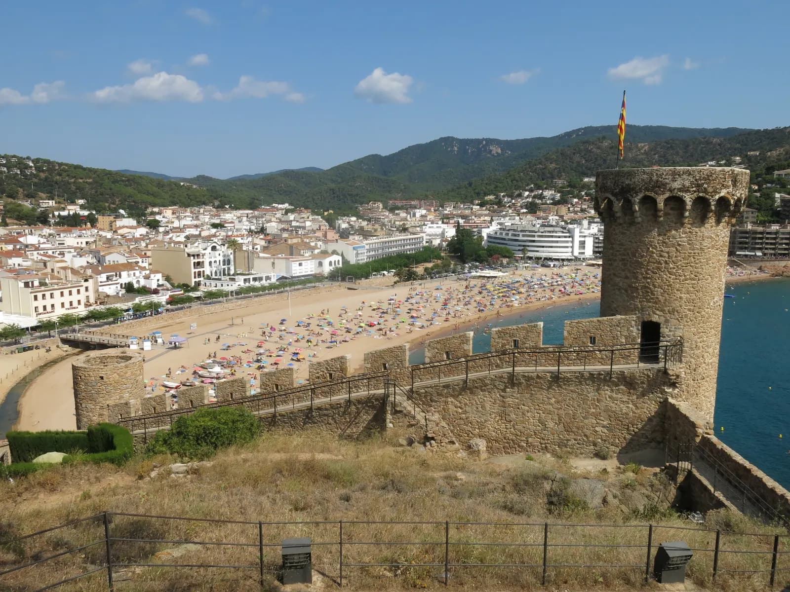 A round medieval tower on the walls of Tossa de Mar's Vila Vella, above the town's main beach and the wooded hills behind.