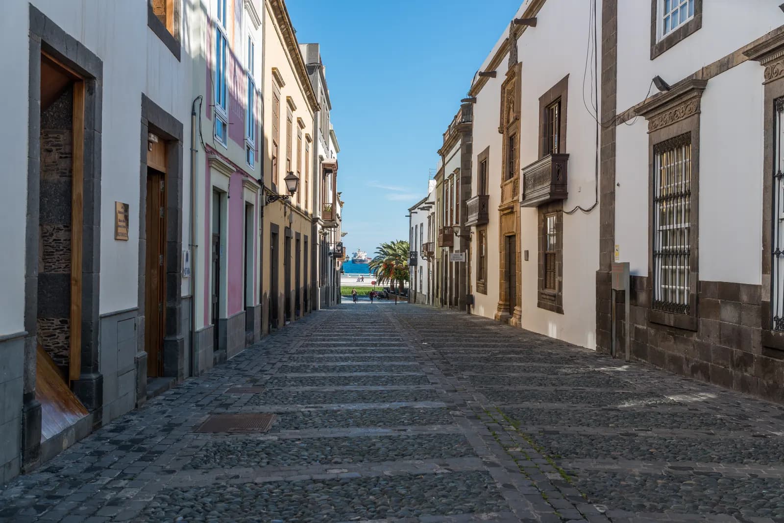 Cobbled street in Vegueta between old houses with stone-framed doorways and a wooden balcony, with palm trees and a glimpse of sea at the far end