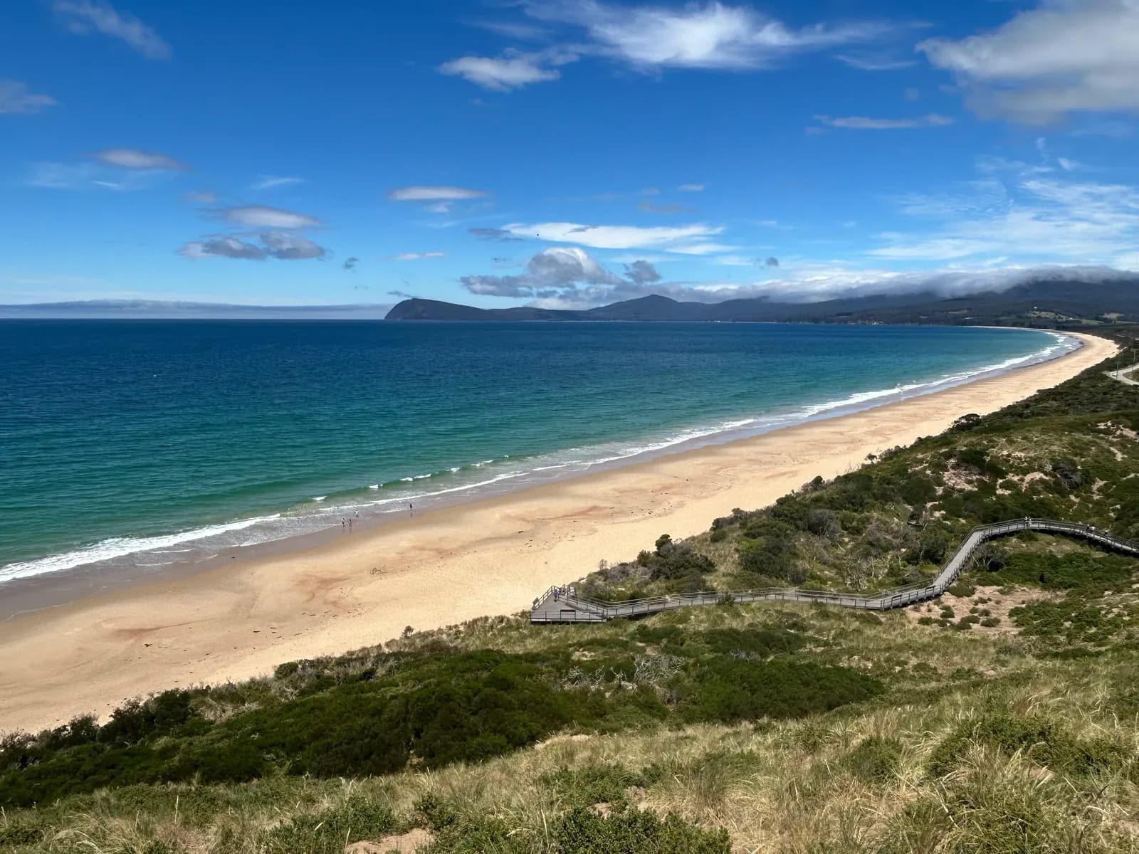 A long sandy beach curving along Adventure Bay, seen from The Neck lookout on Bruny Island, with a timber boardwalk crossing the dunes and hills under low cloud in the distance
