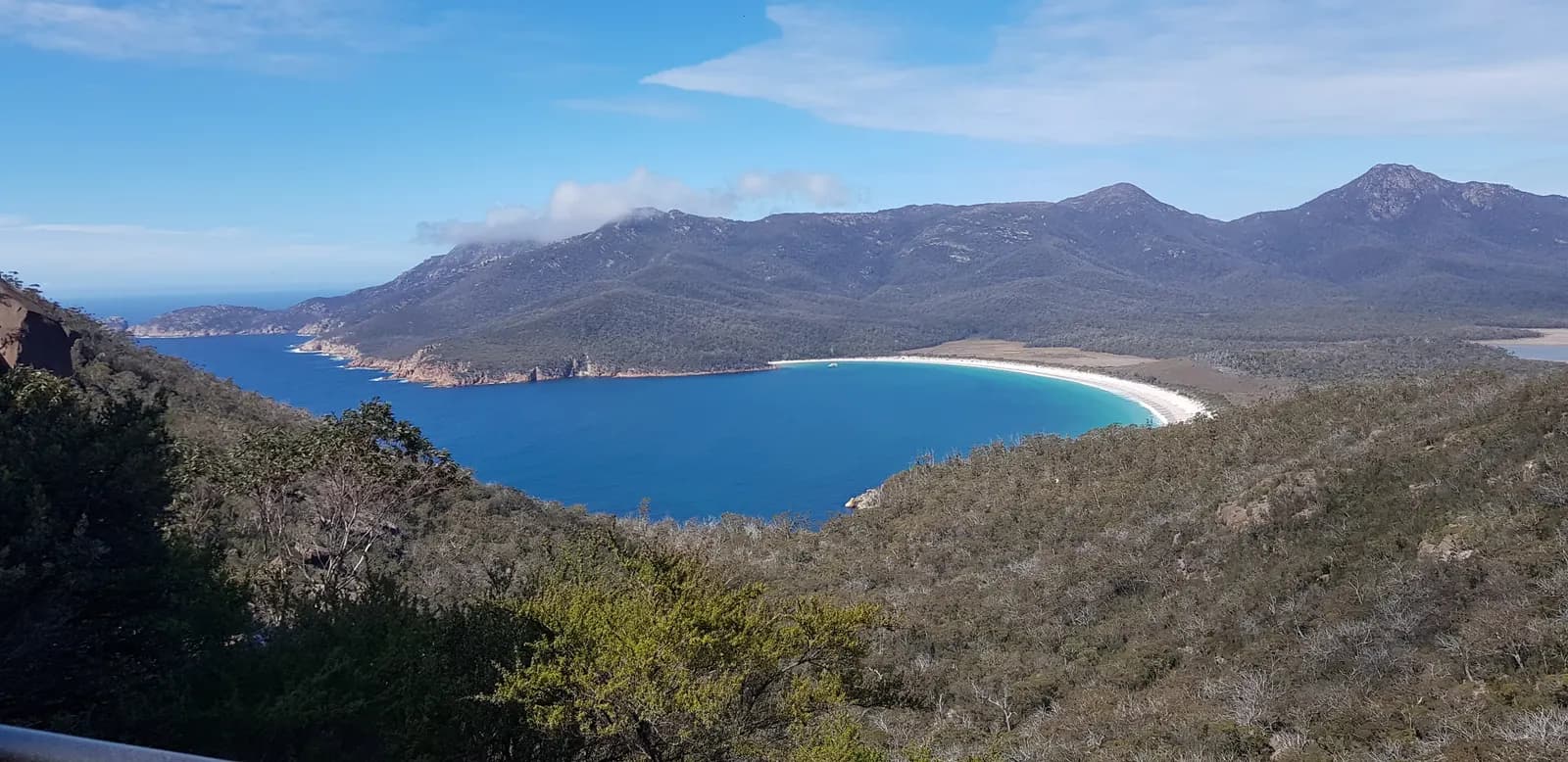 A curved white beach and a blue bay below forested mountains in Freycinet National Park, seen from a lookout