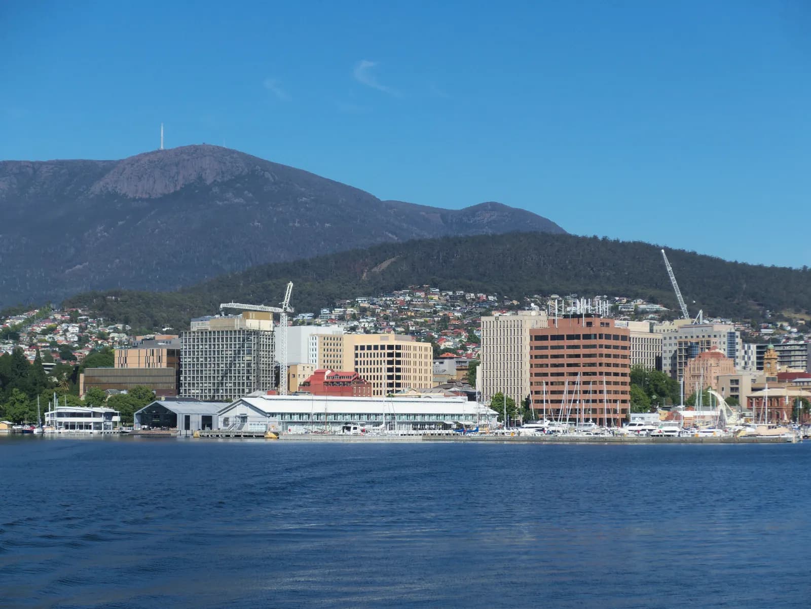 The Hobart city centre and waterfront seen from the water, with wharf sheds, marinas and office buildings, houses on the lower hills and Mount Wellington behind under a clear sky