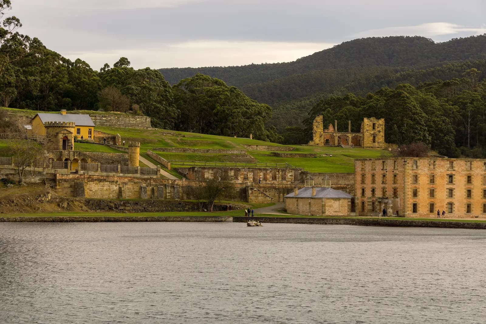 The ruined penitentiary and other sandstone buildings of the Port Arthur historic site seen across the water, with a round stone tower, lawns and forested hills behind
