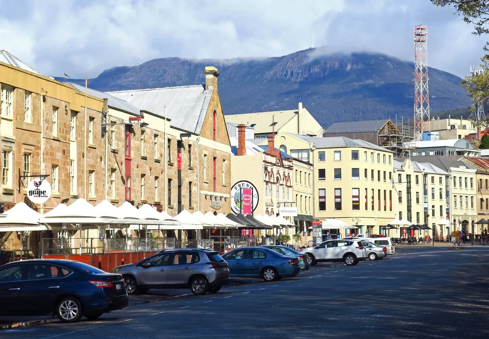 Sandstone warehouses along Salamanca Place in Hobart with outdoor umbrellas and parked cars, and Mount Wellington under cloud behind the rooftops