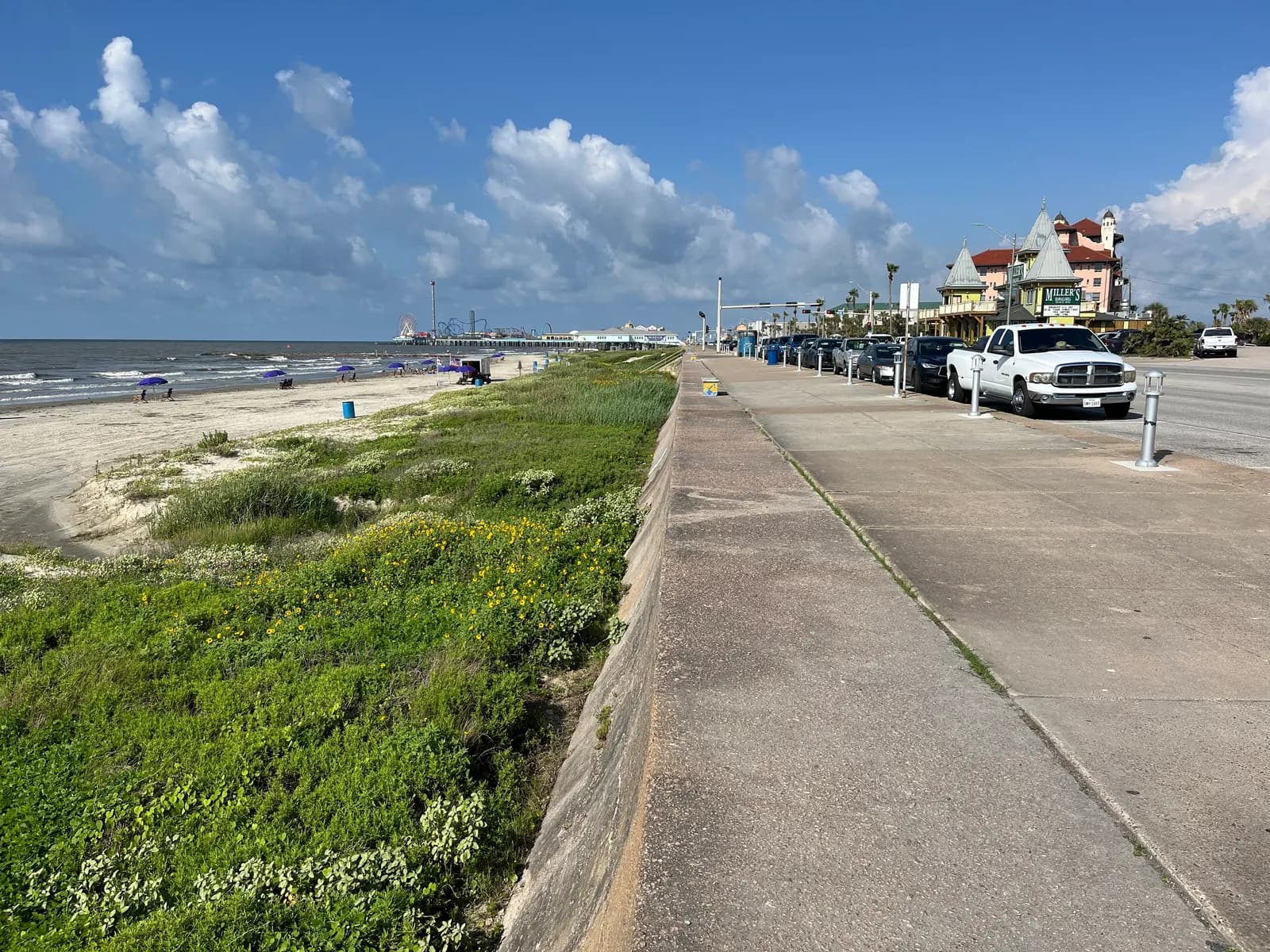 Cars parked along Seawall Boulevard in Galveston, with the seawall walkway, the beach and the Pleasure Pier in the distance