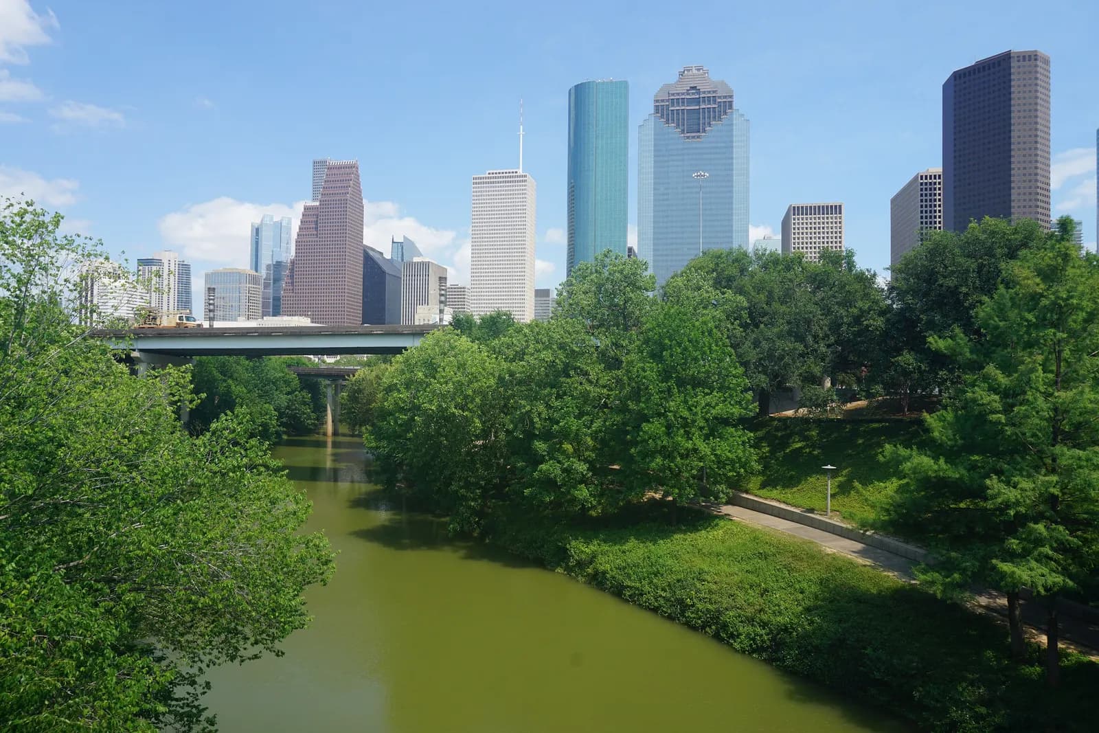 The Downtown Houston skyline above the trees of Buffalo Bayou Park, with the bayou, a footpath and a road bridge in the foreground