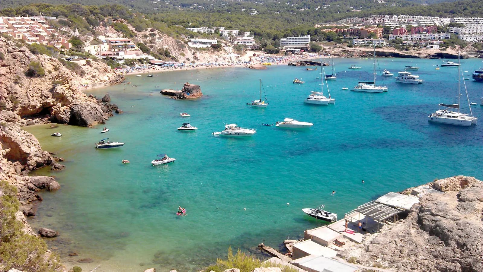 The turquoise bay of Cala Tarida seen from the cliffs, with anchored boats, the beach and holiday apartments on the slopes.