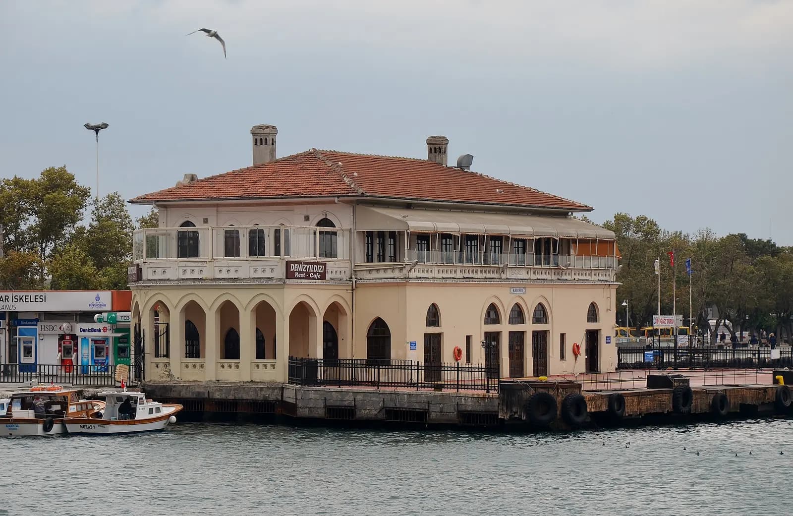 The old ferry pier building of Kadıköy, with arched windows and a tiled roof, on the water with small boats moored beside it