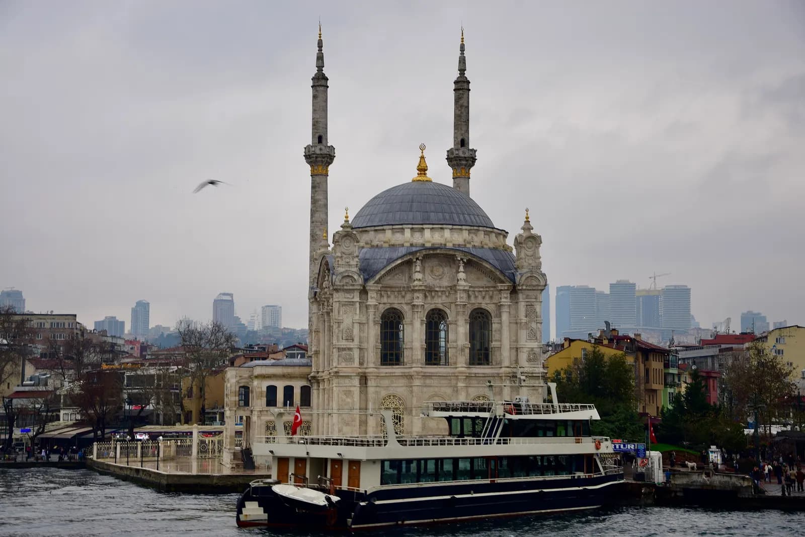 The Ortaköy Mosque on the shore of the Bosphorus with a moored boat in front and high-rise towers on the skyline behind