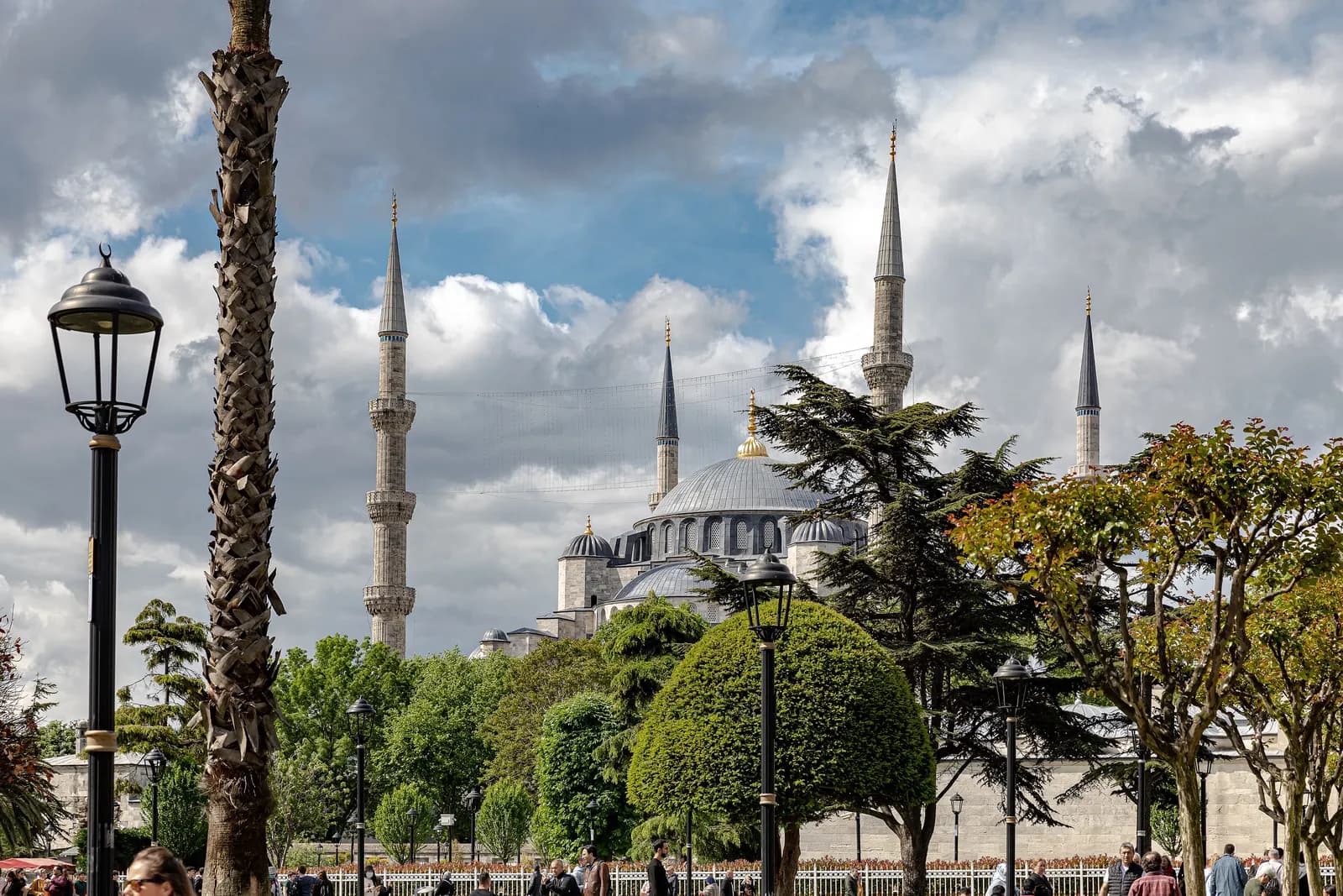 The domes and minarets of the Blue Mosque above the trees and street lamps of Sultanahmet Square under a clouded sky