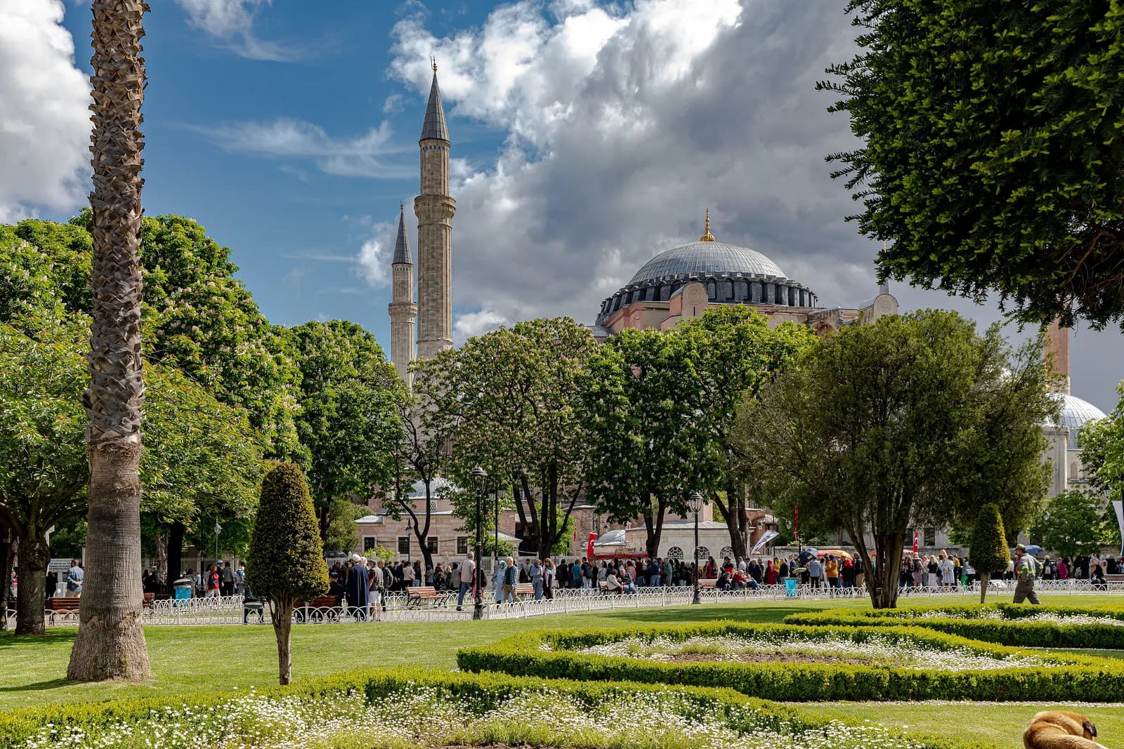 The dome and minarets of Hagia Sophia behind the trees and flower beds of the Sultanahmet park, with visitors along the railings